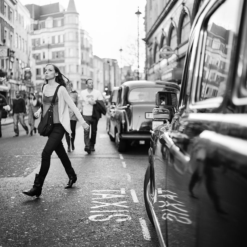 Black and white street photograph of a woman crossing the road as London black cabs wait nearby